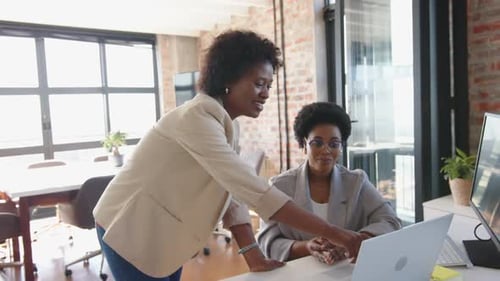 Discussing project, two businesswomen collaborating at desk with computer in office