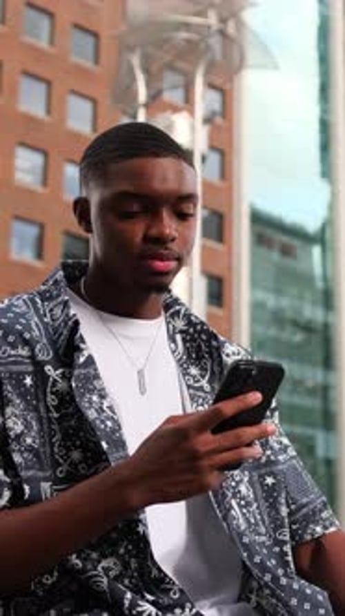 An African American Man Stands on a City Sidewalk and Looks at His Smartphone