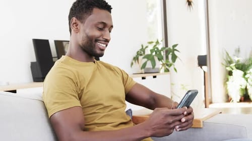 Happy african american man using smartphone sitting on couch in living room, in slow motion