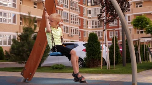 A Little Happy Boy Rides on a Swing on a Modern Playground in the Courtyard