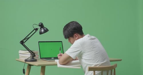 Asiatischer Mann schaut auf einen Greenscreen-Laptop und schreibt auf das Buch, während er auf dem Greenscreen lernt