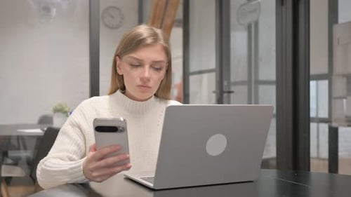 Woman Uses Phone While Working on Laptop