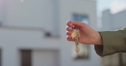 Closeup of a Key with a Houseshaped Keychain in Hand In the Background is a House Real Estate
