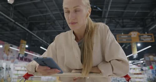 Young Lady Using Smartphone and Looking at Goods in Shopping Cart Walking in Supermarket Wearing