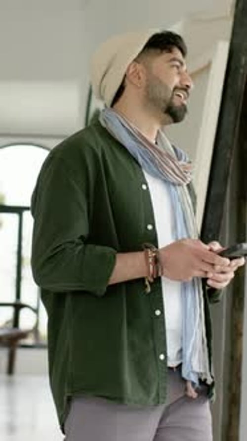 Vidéo verticale : jetant un coup d'œil depuis la toile d'un studio lumineux, un homme portant un bonnet et un foulard fait défiler le téléphone
