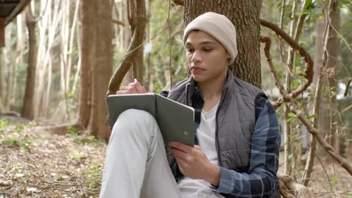 Young man in forest writing in notebook, reflecting on nature's beauty