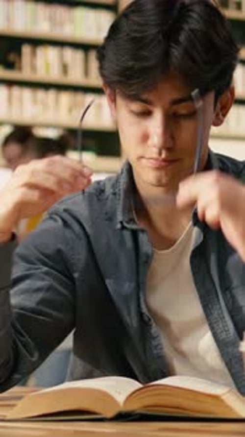 Vertical Video Tired Asian Student Leaning Over Open Book Between Tall Stacks in Library