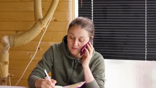 Femme parlant au téléphone tout en regardant vers le bas en écrivant dans un cahier, inclinée vers le bas
