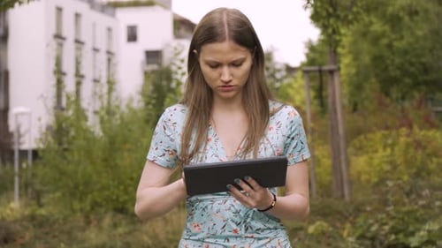 A Young Caucasian Woman Works on a Tablet As She Walks in a Suburban Area with an Apartment Complex