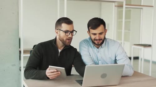 Two male office workers sitting at a laptop and a tablet doing work together