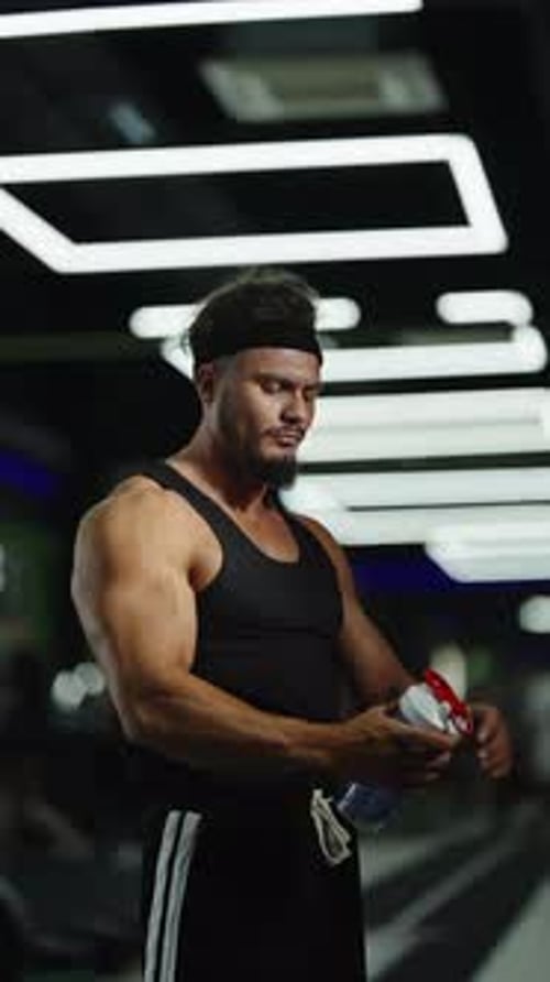 Confident Muscular Athlete in a Modern Gym Wearing a Black Tank Top and Headband Holding a Shaker