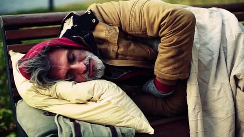 Homeless Beggar Man with a Pillow Lying on Bench Outdoors in City,
