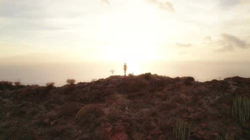 Silhouette of a person in orange outfit with arms raised facing the sunrise over the ocean, standing