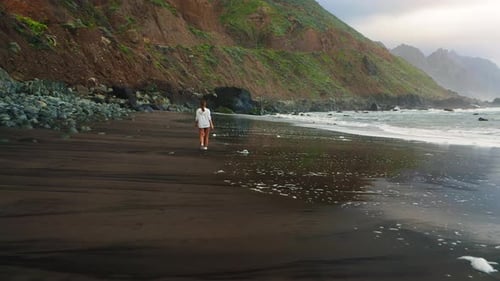 Young Woman Tourist Walks Along the Black Volcanic Sand Beach at Sunset