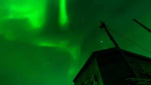 Otherworldly aurora borealis flare in night sky above boat house, nightlapse