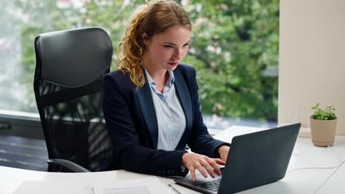 in a Modern Office with a Large Window a Beautiful Worker in a Blue Shirt with a Laptop Looks at the