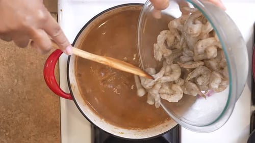 Adding raw shrimp to the gumbo roux mix and sausage simmering in a pot on the stove - overhead view