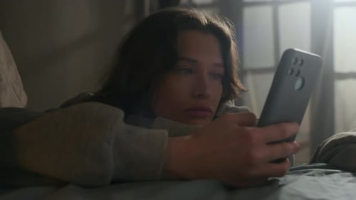 Bored Girl Doomscrolling on Smartphone while Lying in Bed at Home