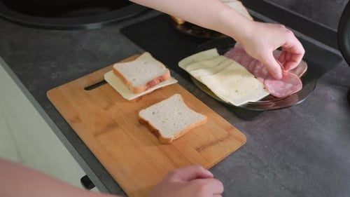 Hand Assembling Sandwiches Using Slices of White Bread Cheese and Deli Meats on Wooden Cutting Board