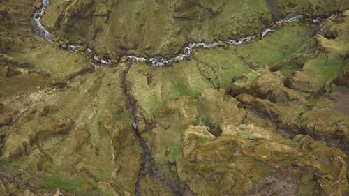 Aerial shot of a winding stream cutting through a rugged, moss-covered landscape in Iceland, with ro