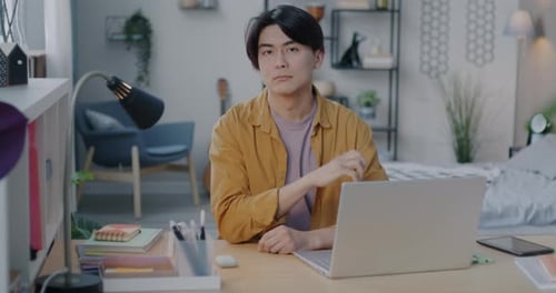 Slow Motion Portrait of Young Asian Man Sitting at Desk with Laptop Smiling Looking at Camera