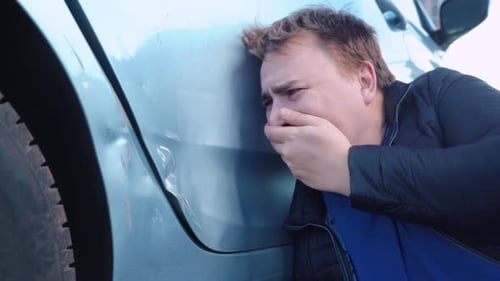 Broken automobile in parking lot. Guy is crying hysterically because of dented car door, beating it