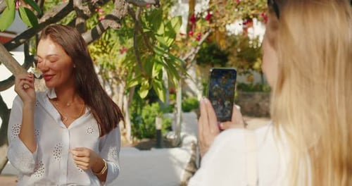 Woman holding a flower near her face while posing, being photographed outdoors in a tropical garden.