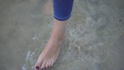 Barefoot woman standing in sea water moving on sandy beach in slow motion
