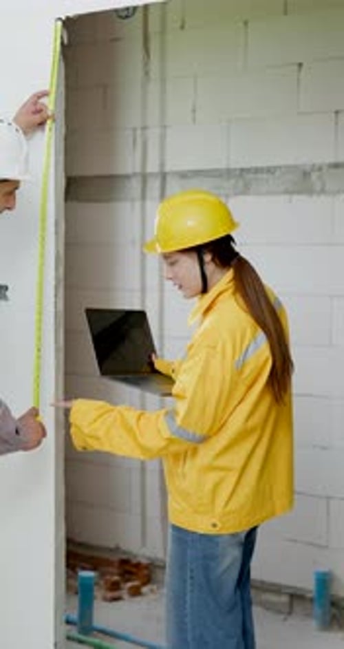 Asian woman engineer reviews laptop by unfinished wall while male contractor measures surface,