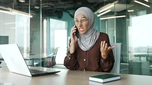Irritated arguing young muslim businesswoman in hijab yelling shouting on smartphone at office Angry