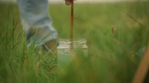 Artist Rinsing Brush in a Jar of Water Amidst Green Grass Outdoors