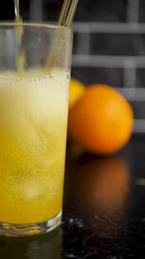 Vertical video: citrus drink pouring into glass cup against oranges and black wall. Carbonated orang