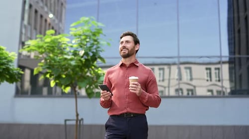 Confident male entrepreneur in red shirt enjoying coffee while using phone outside modern city offic
