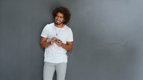 Man Leaning Against A Grey Wall Using Mobile Phone. Portrait Of A Happy Guy Texting With Cellphon...