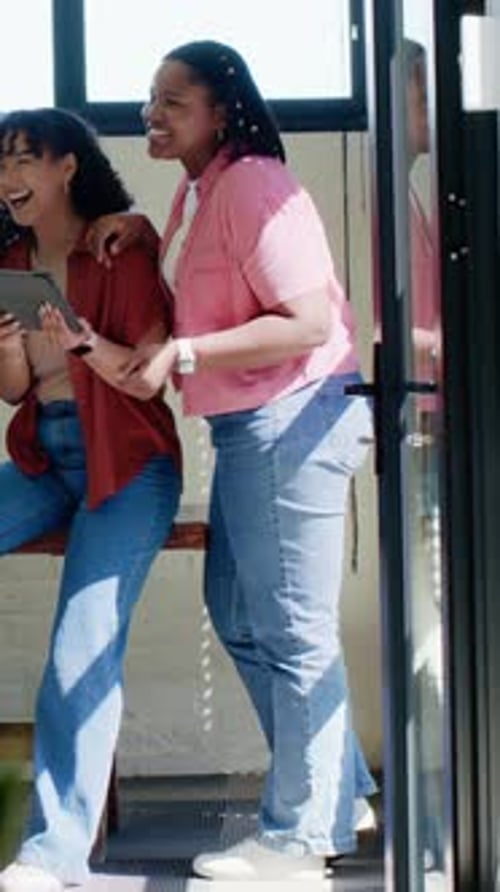 Vidéo verticale : femme assise riant tenant une tablette jouant à un clip, amie faisant des gestes dans une pièce lumineuse