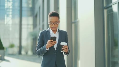 business black woman walking on city street downtown with mobile phone and cup coffee. African