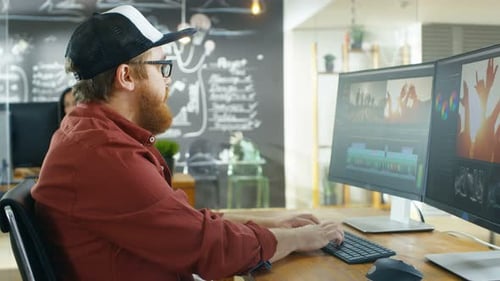 In der Kreativagentur arbeitet Videograf mit Kappe an einem PC mit zwei Monitoren, er ist Doi