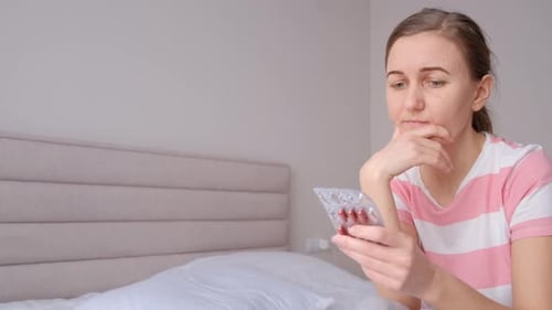 Upset Young Woman Sitting on Bed Holding Pills and Suffering From Headache Pain Chronic Fatigue