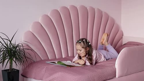 The concept of childhood and leisure. A little girl is lying on the bed looking at the book