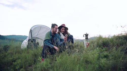 Young couple enjoying nature sitting outside their tent shelter with smartphone