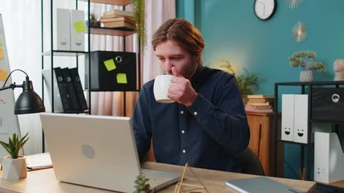Geschäftsmann arbeitet am Laptop und trinkt eine Tasse warmen heißen Kaffee, sitzt am Tisch, am Arbeitsplatz, im Heimbüro