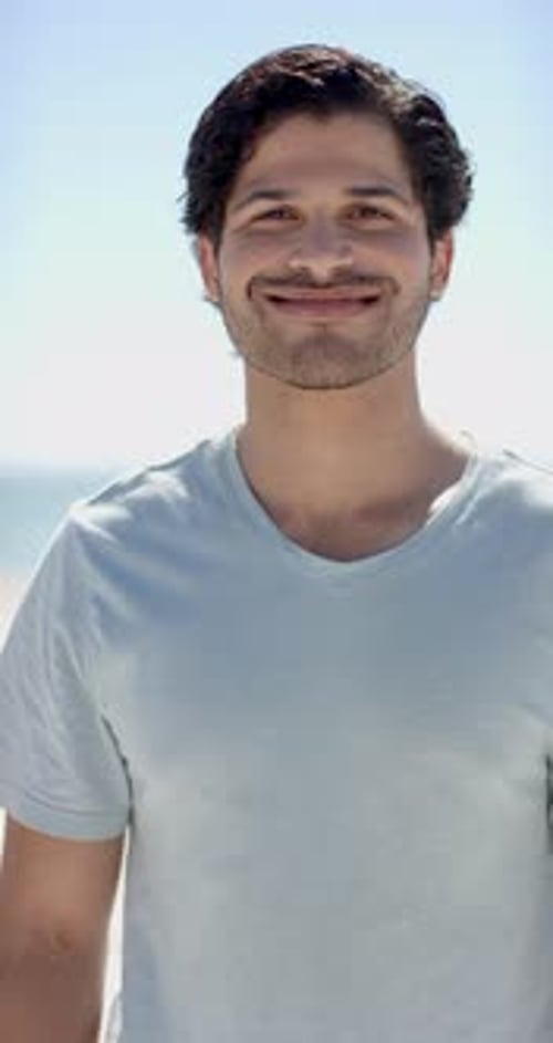 Vertical video: Smiling man enjoying sunny day at beach, wearing casual t-shirt