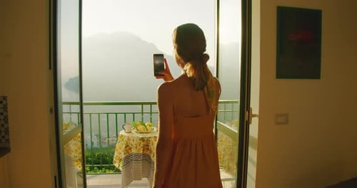 Person captures a sunny mountain view from a balcony with a smartphone. Woman gazes out towards a se