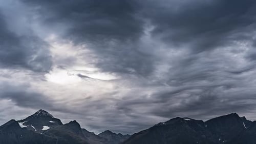 Stormy clouds backlit by the sun pass in the sky above the dark mountains.