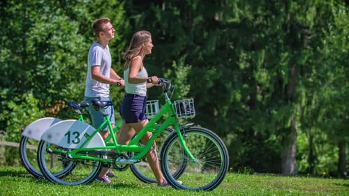 Young couple walks with bikes in a park and approaches a landmark statue