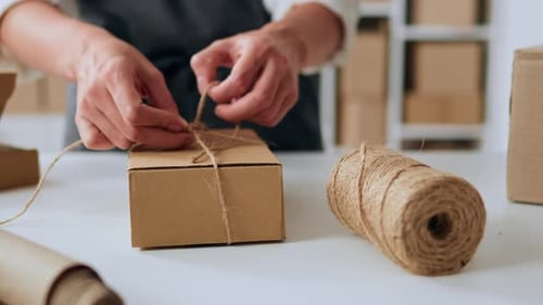 A Packer In A Warehouse Is Tying Up A Gift For A Customer. Craft Paper Packaging By Warehouse Worker