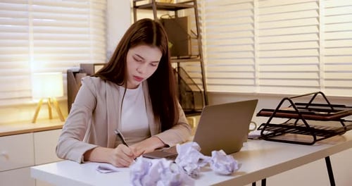 Asiatische Mitarbeiterin sitzt am Schreibtisch und wirft vor Stress Papierball, umgeben von Laptop und