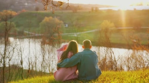 Young people sitting on grass and looking at sunset near the river.