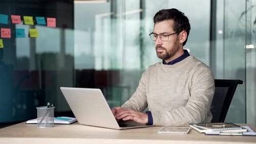 Focused mature businessman working on laptop at desk in office, wearing casual sweater and glasses.