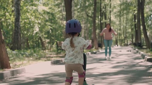 Mom Teaches Daughter to Ride a Scooter in the Park in Summer